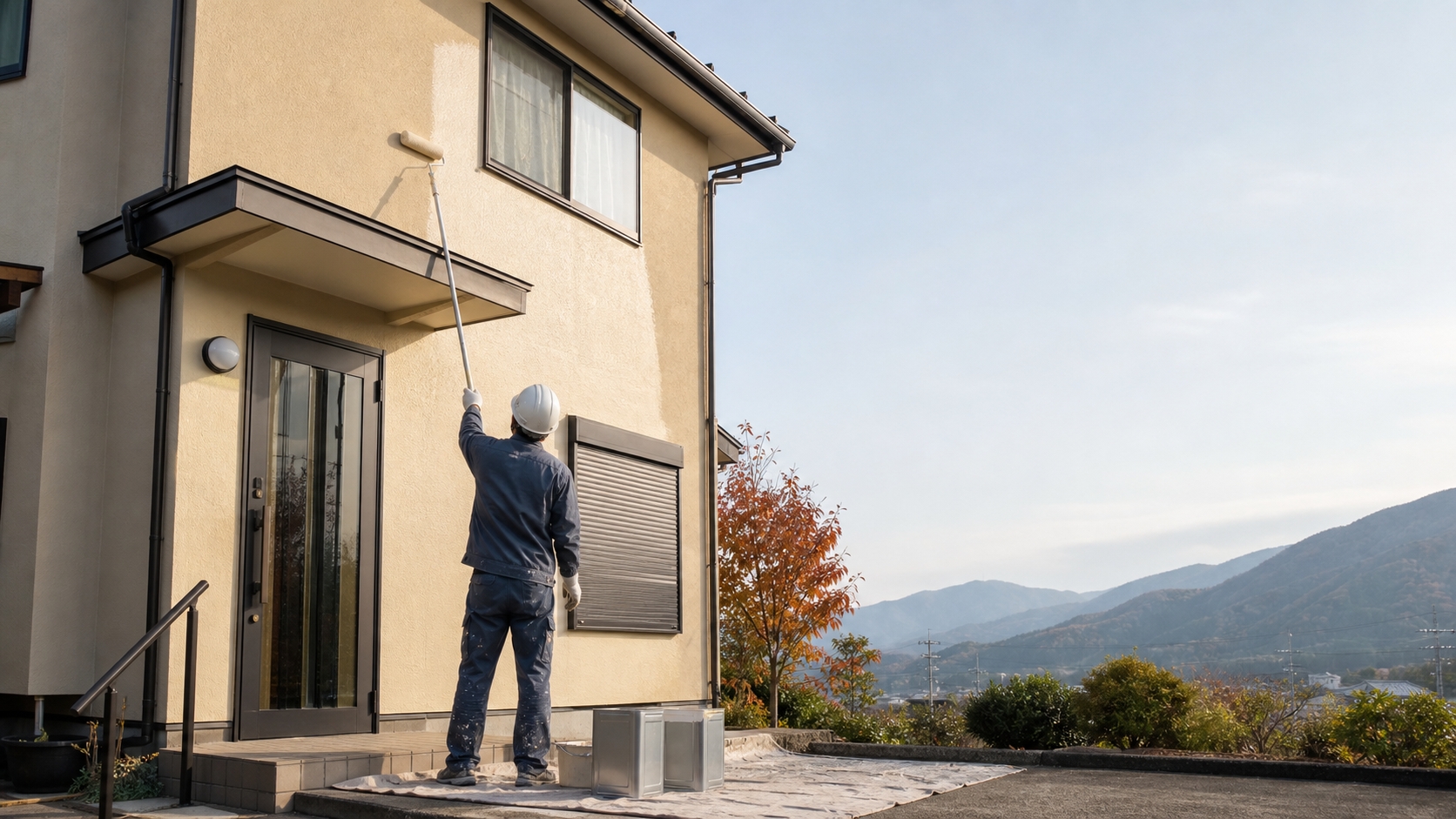 晩秋の晴れた日、山並みを背に戸建て住宅の外壁をローラーで塗装する職人
